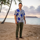 A smiling man wearing a blue and white tropical floral print shirt stands on a sandy beach at sunset, with a tropical island and palm trees in the background.