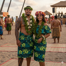 A couple standing on a tropical beach wearing matching custom-made island attire. The man is in a traditional sarong and the woman in an off-the-shoulder dress, both crafted from green Hibiscus, Plumeria, and Monstera leaf patterned polyester fabric. They are adorned with floral leis and headpieces amidst a cultural outdoor celebration.