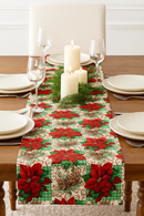 A festive table setting featuring a Poinsettia Pine Holiday Plaid table runner on a wooden table. The runner has a pattern of red poinsettias, pinecones, and green holly on a green and white plaid background. The table is set with white plates, wine glasses, and a centerpiece of white pillar candles and evergreen branches.
