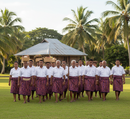 A group of Tongan men walking across a lush green lawn in front of a traditional fale (house) with a thatched roof and palm trees. They are wearing formal white short-sleeved shirts and matching purple Traditional Tongan Geometric Design Peachskin Fabric wraps around their waists, known as tupenu. Some men are carrying woven baskets on their heads and one holds a wooden staff, capturing a moment of cultural ceremony and tradition.